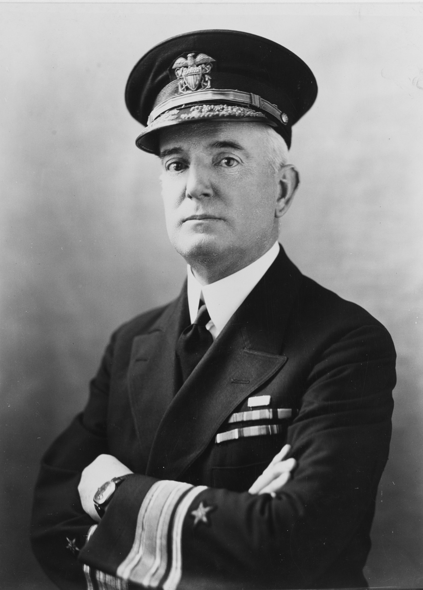 A headshot of Rear Admiral William A. Moffett wearing his dark, formal Navy uniform and admirals hat with his arms crossed.