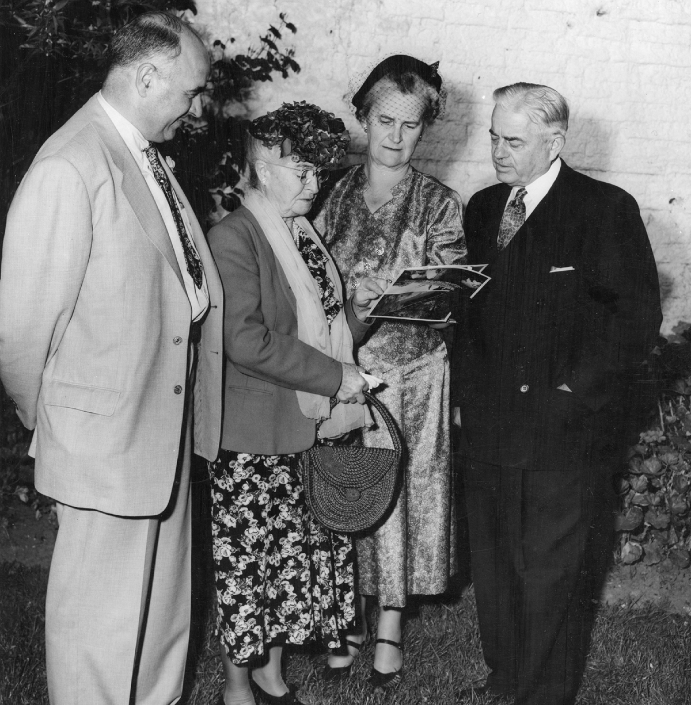 Laura Whipple wearing a dress, coat, and hat. She is standing beside 2 men in suits and a woman wearing a dress and hat. They are all looking at a photo.