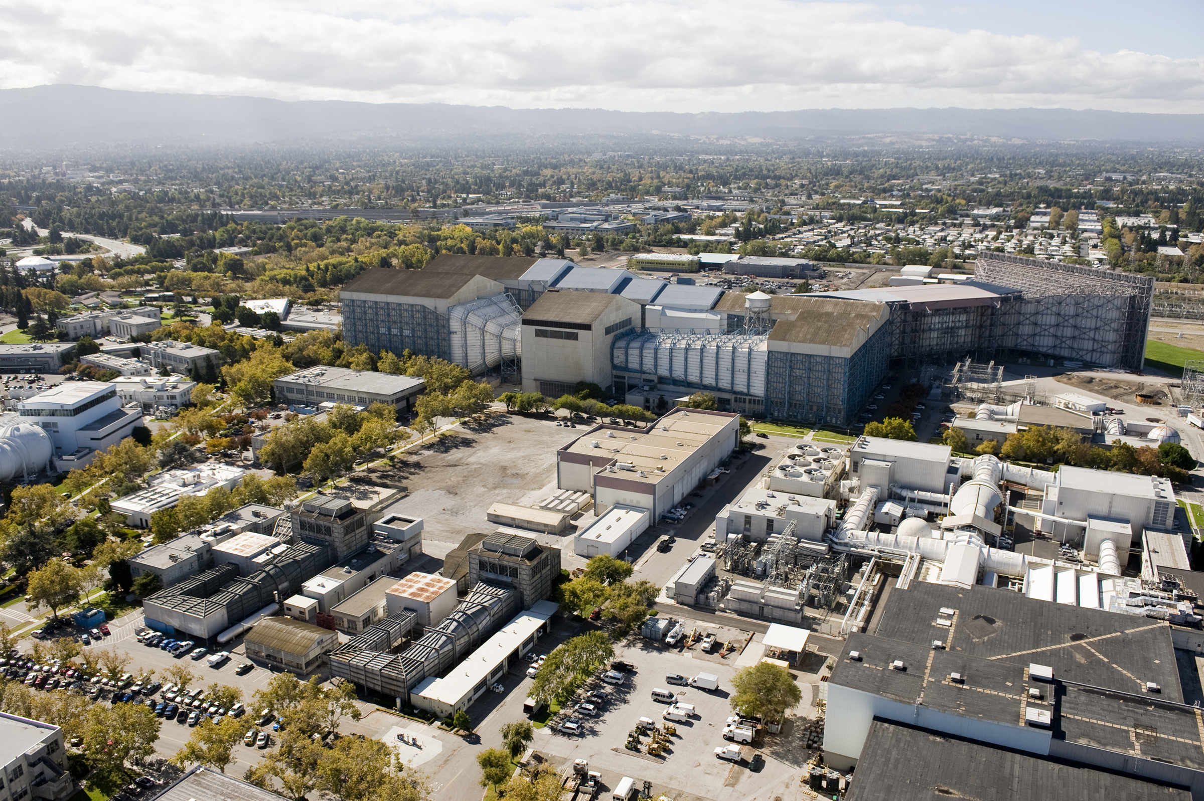 A birds-eye view of large wind tunnel buildings with long, curved chambers connected to huge industrial structures with flat and peaked roofs.