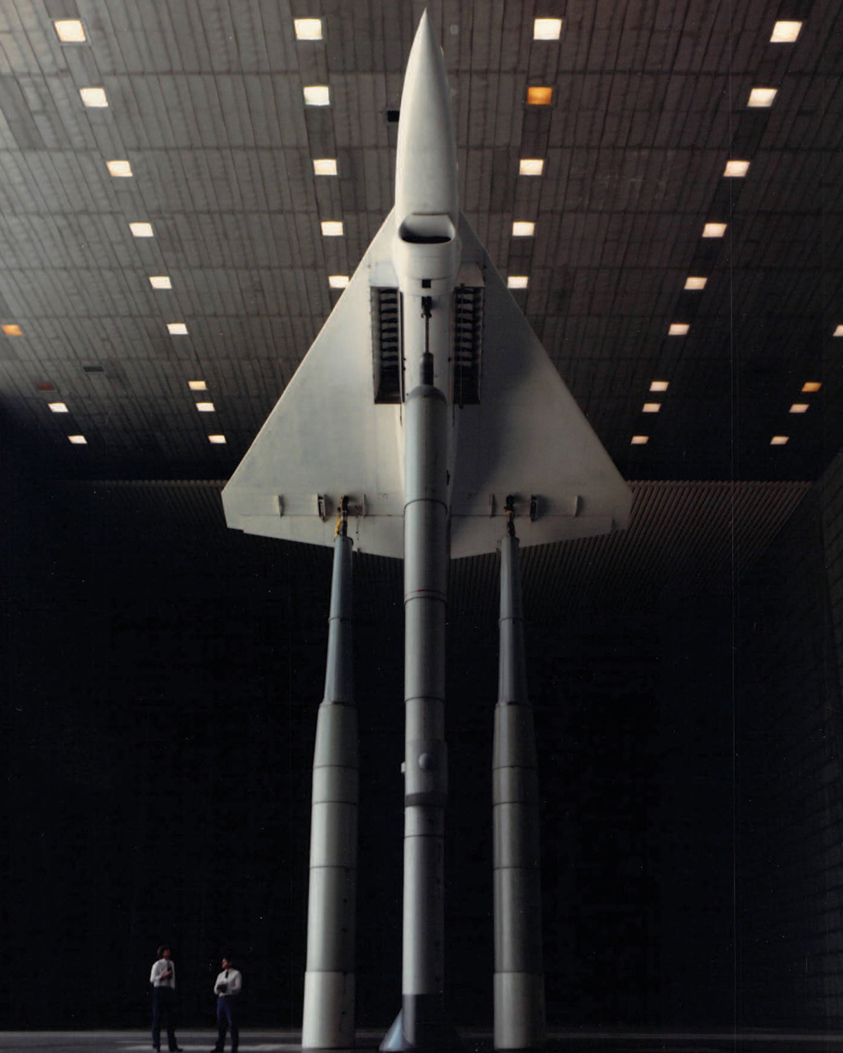 Two men stand below an aircraft raised above the floor on tall columns, with a triangular body and sharp nose cone pointed to the wind tunnel ceiling.