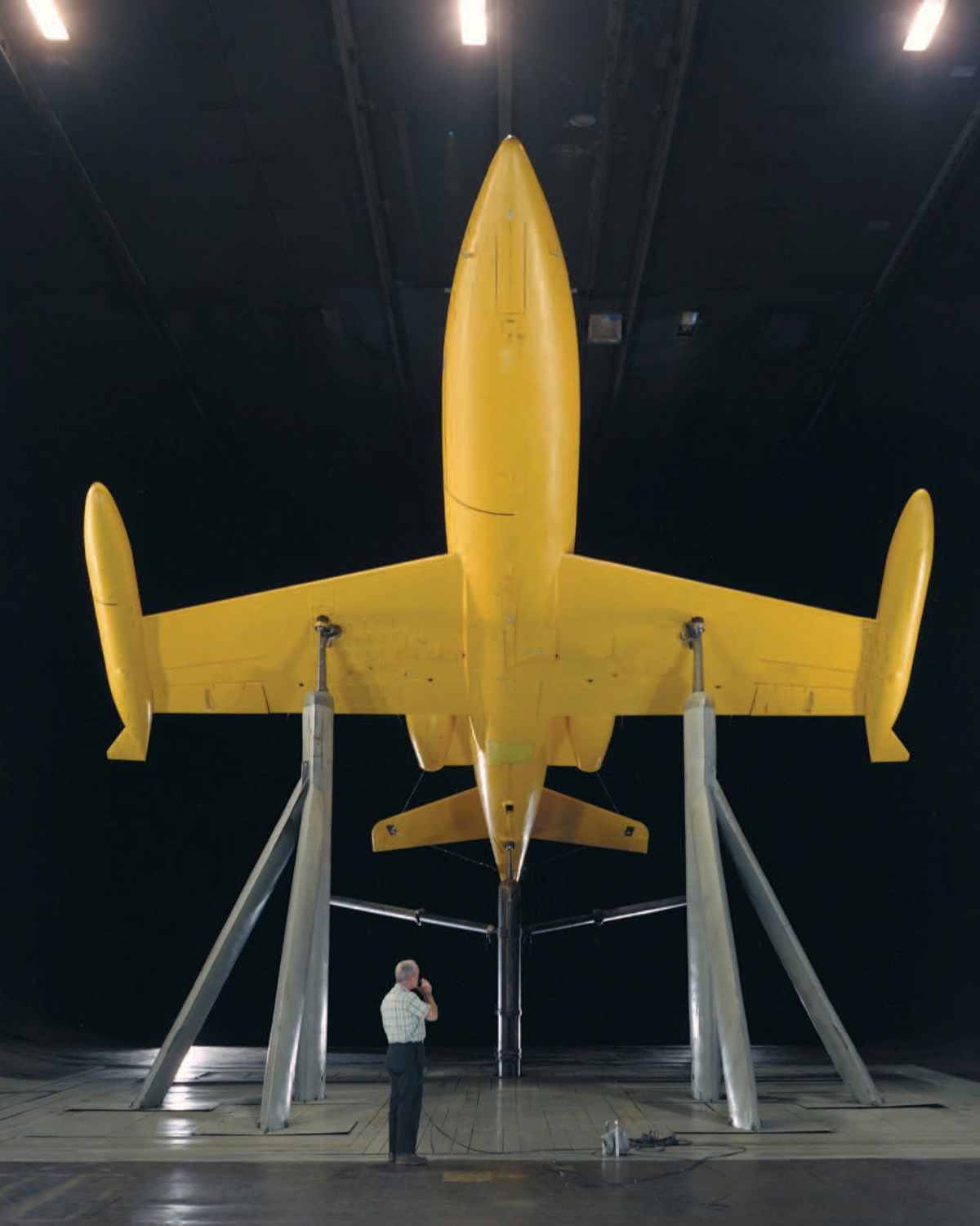 A sleek, yellow jet with its nose cone pointed up toward the wind tunnel ceiling is studied by a man standing beneath its aerodynamic wings.