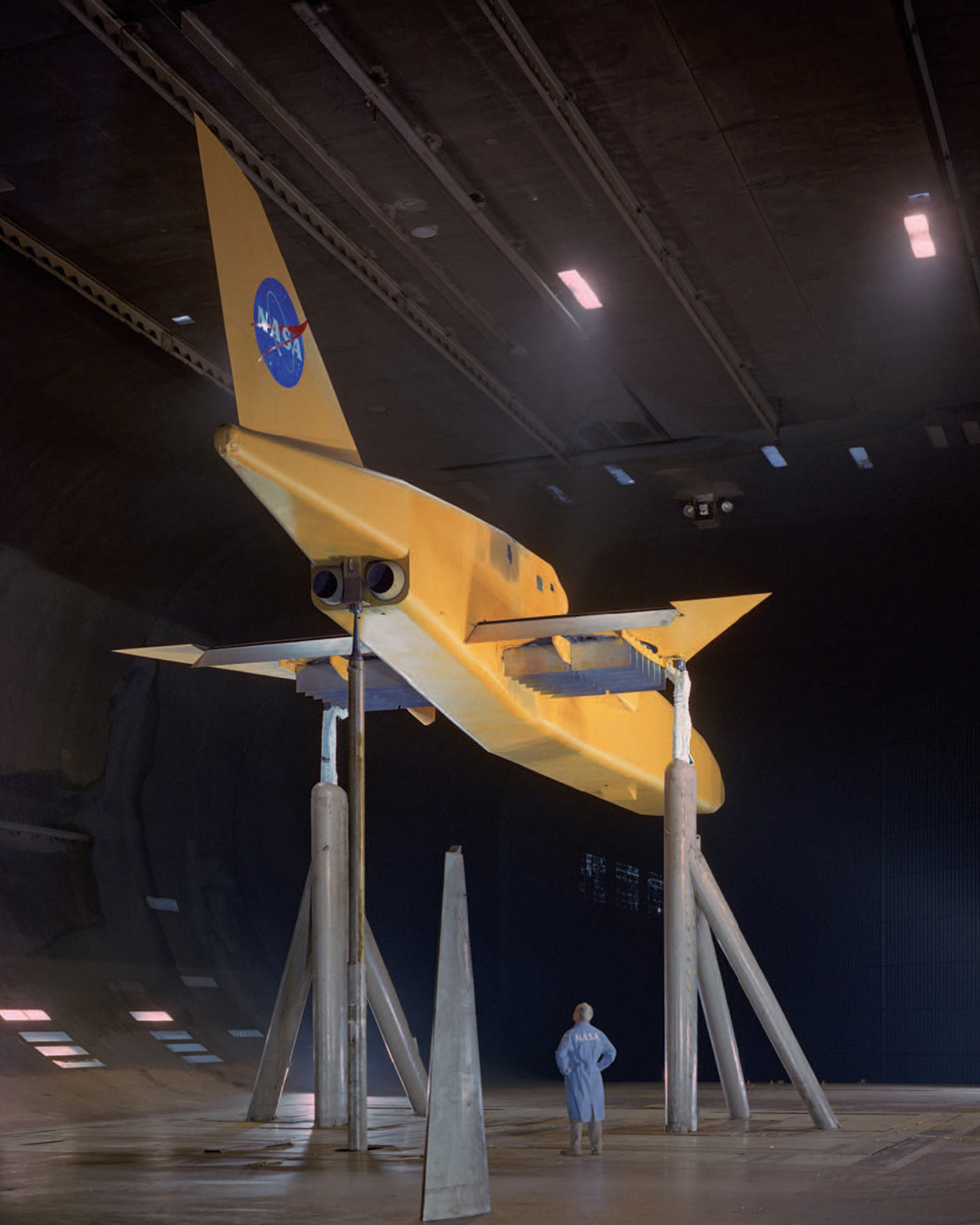 A man looks up at a yellow aircraft with short wings and a tail with NASA’s logo. The plane is raised above the floor on tall columns inside a wind tunnel.