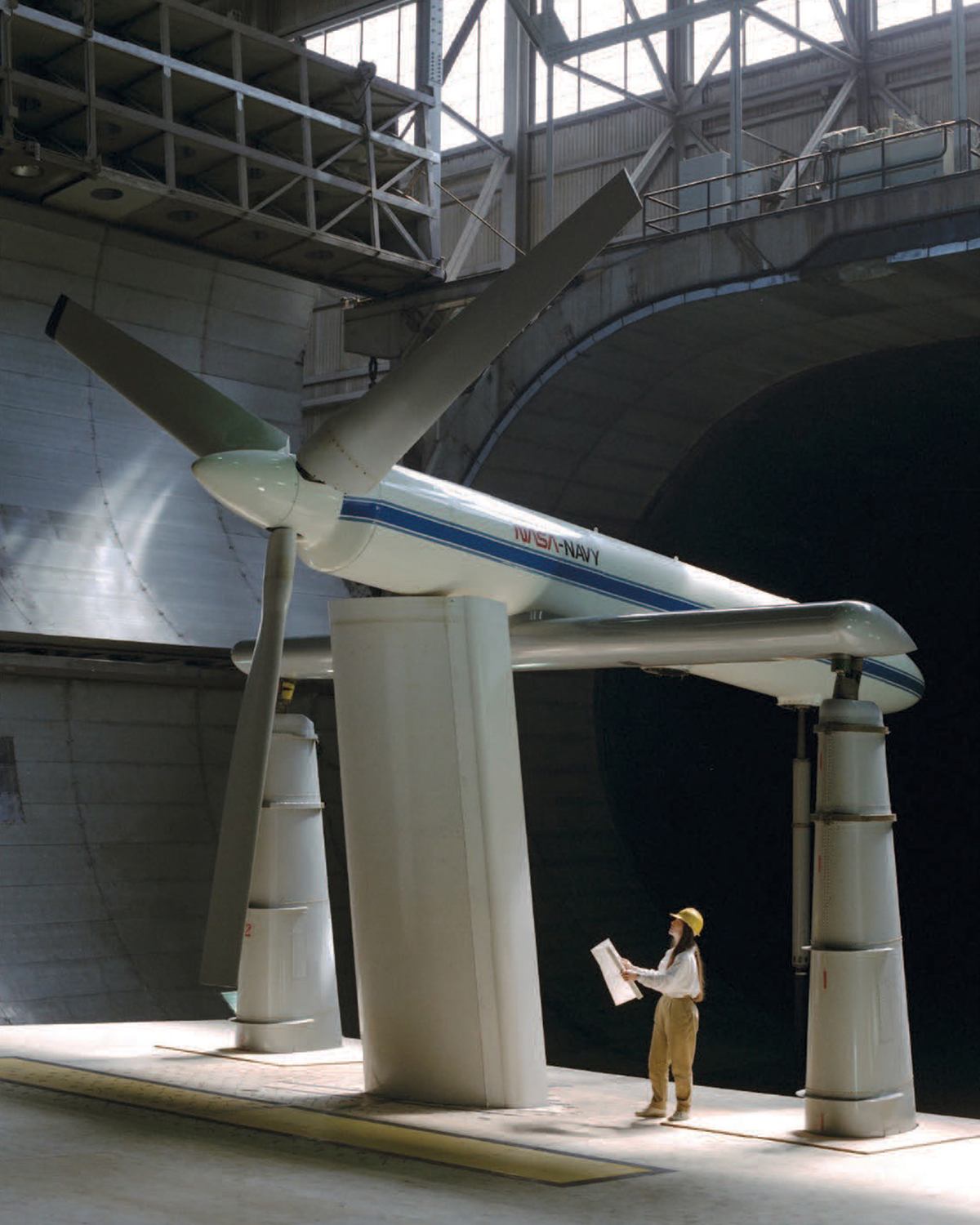 A woman wearing a hard hat and holding a large drawing looks up at a huge propeller with 3 long gray blades that is being tested inside a wind tunnel.