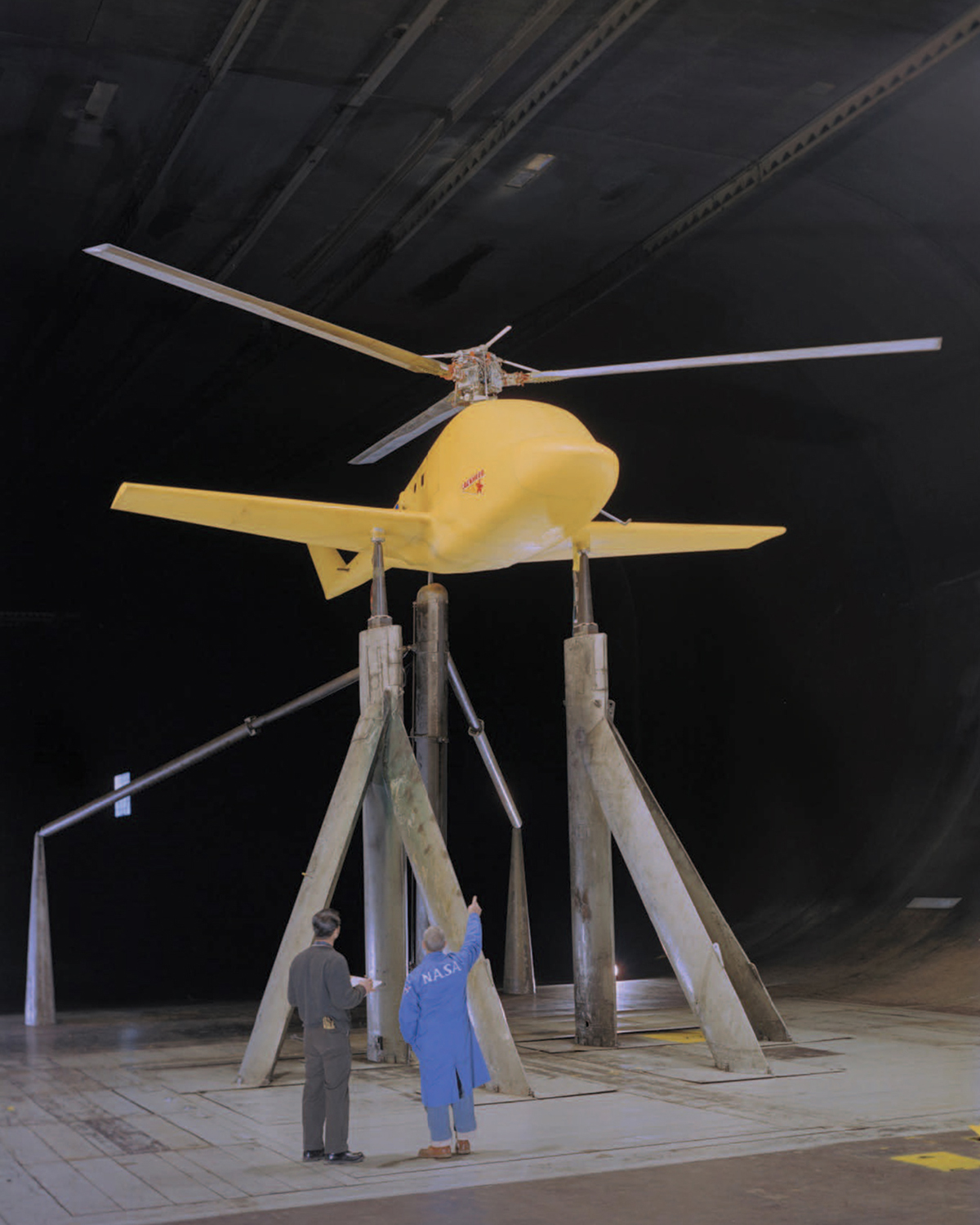 Two men, with their backs to the camera, look up at a yellow helicopter with wings that is raised above the floor on tall columns inside a wind tunnel.