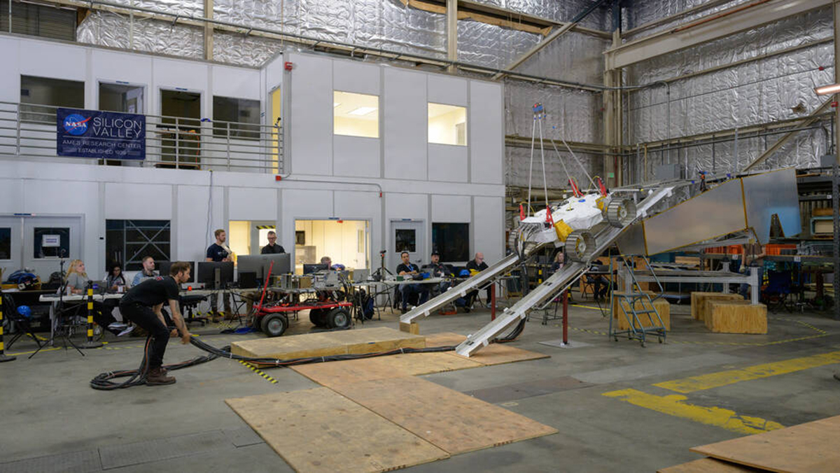 A man holding a long cable guides a white, flat, square-shaped vehicle down 2 metal tracks from a raised platform to the floor of a large research lab.