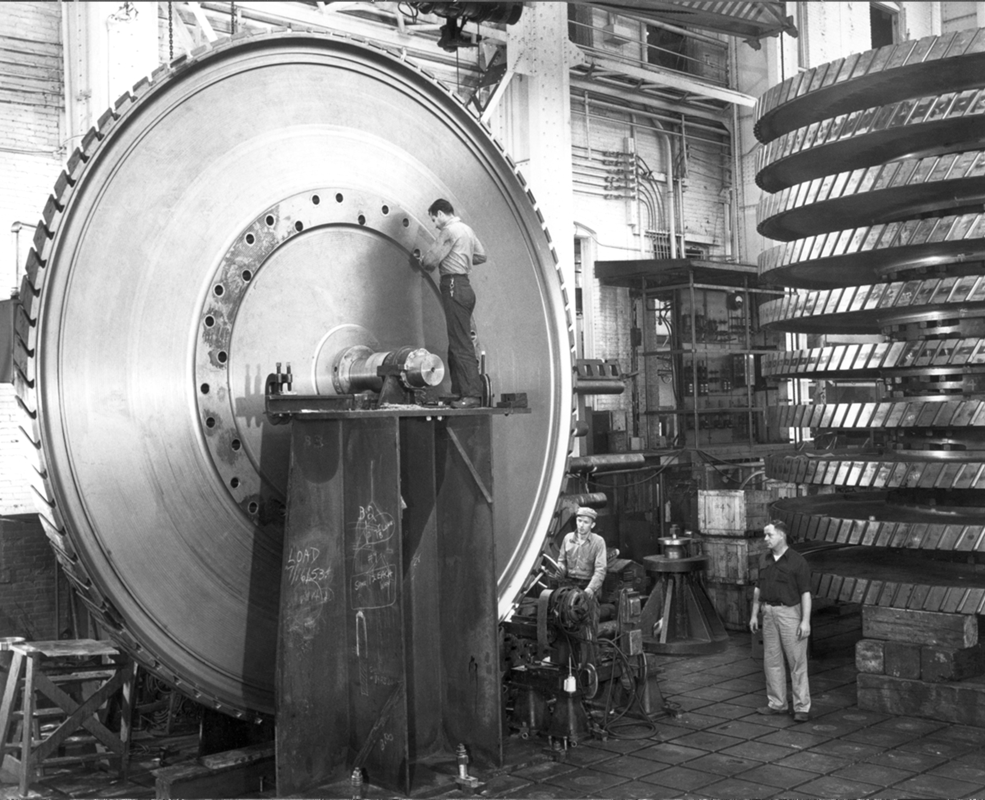 A man repairs a huge, circular gear, while standing atop a tall metal platform attached to the big machine. 2 men stand beside a stack of 10 huge gears.
