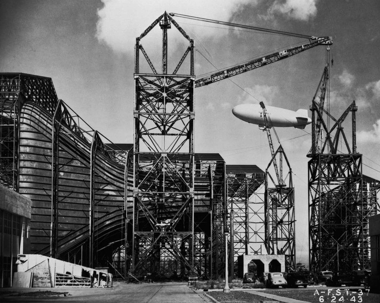 Three steel construction cranes stand beside the curved metal exterior of an under construction wind tunnel. A nearby blimp ascends into the sky.
