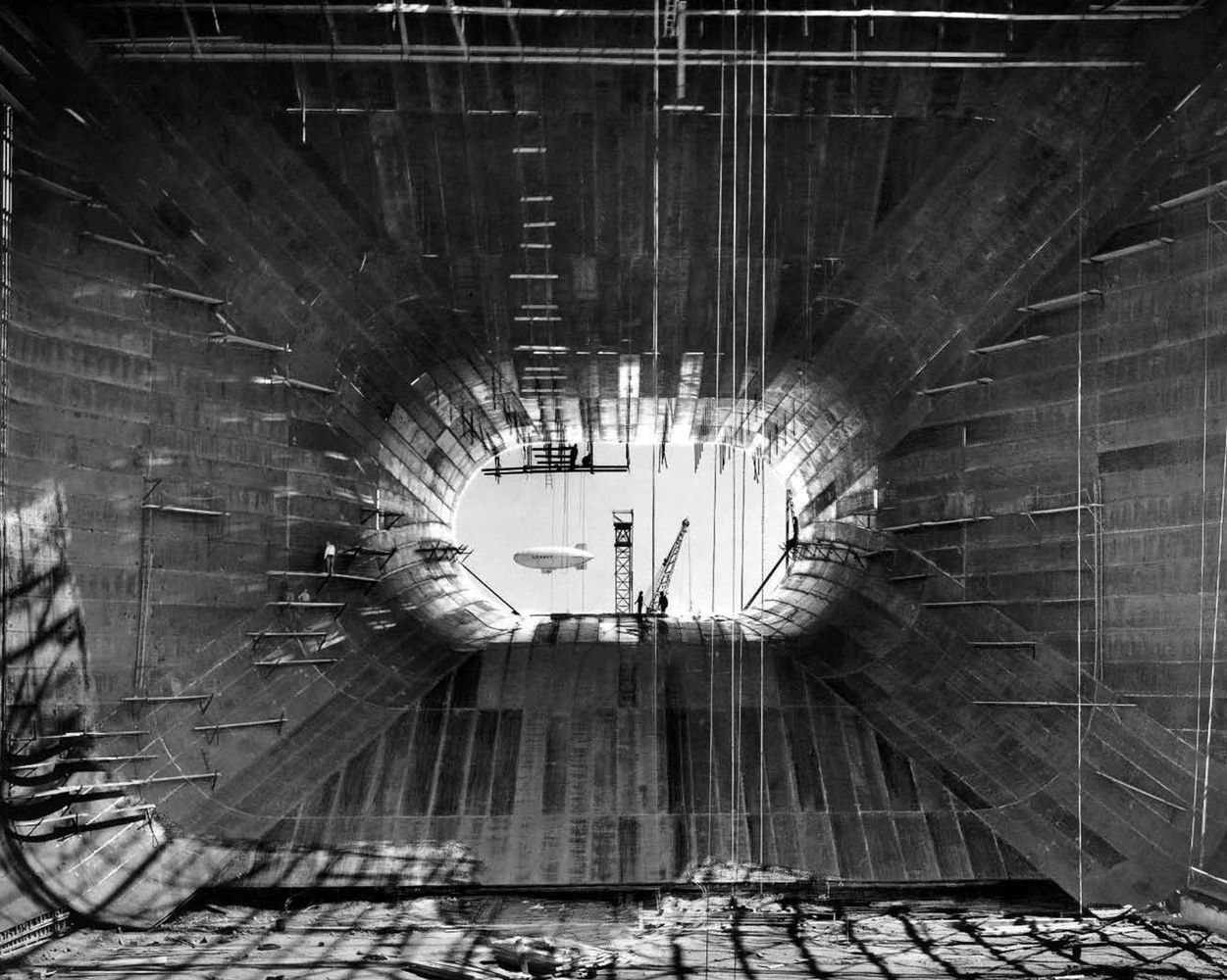 A dark section of a large wind tunnel with an oval opening at its center that reveals a blimp flying in the distance and a construction crane.