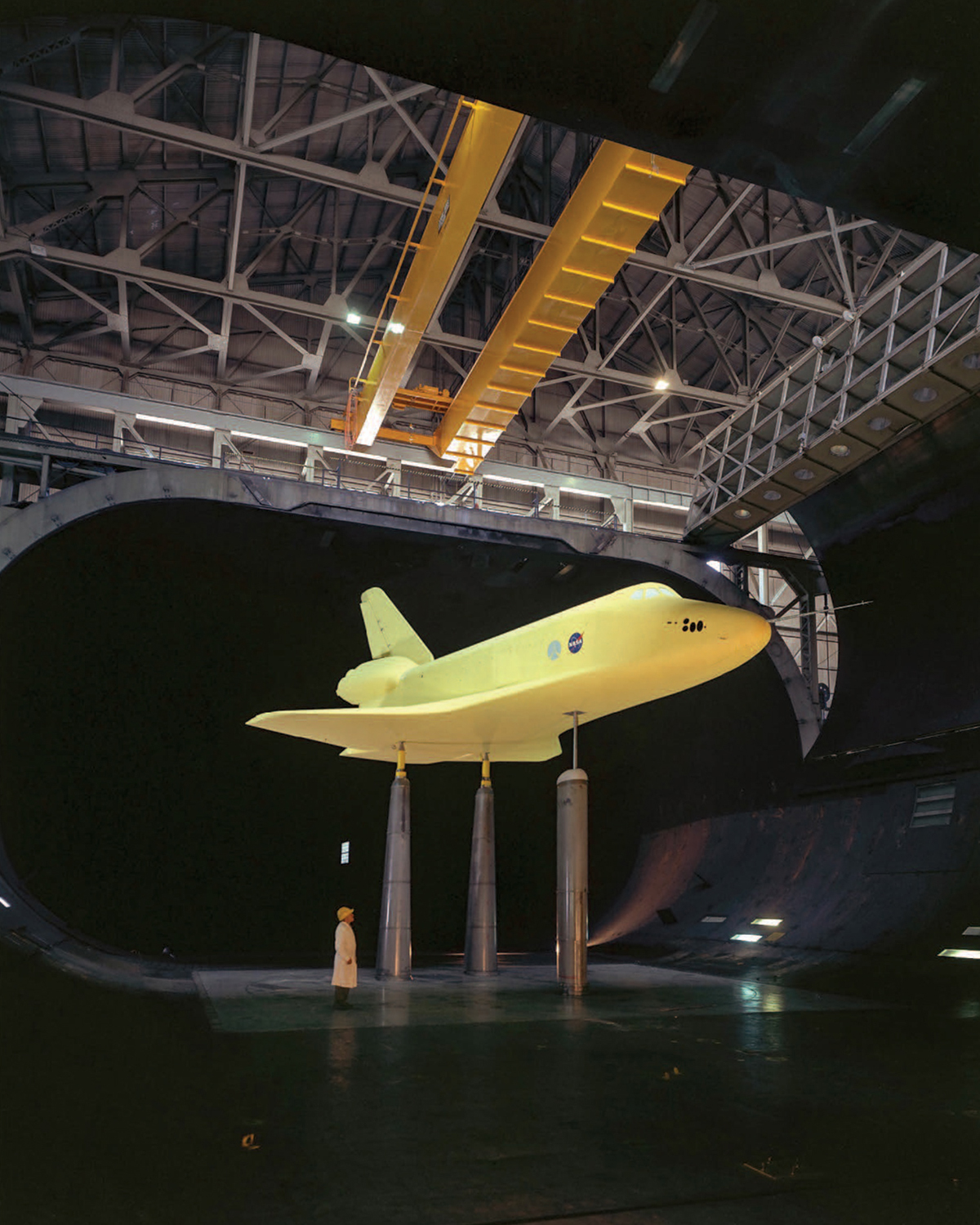 A man wearing a hard hat looks up at a very large scale model of a space shuttle, which is raised above the floor on tall columns inside a wind tunnel.