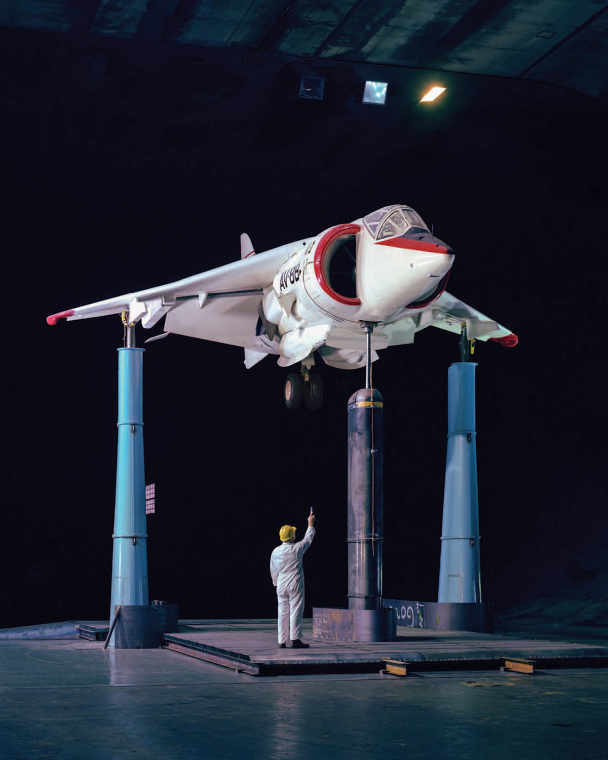 A man in a hard hat with his back to the camera points up at a white fighter jet, which is raised above the floor on tall columns inside a wind tunnel.
