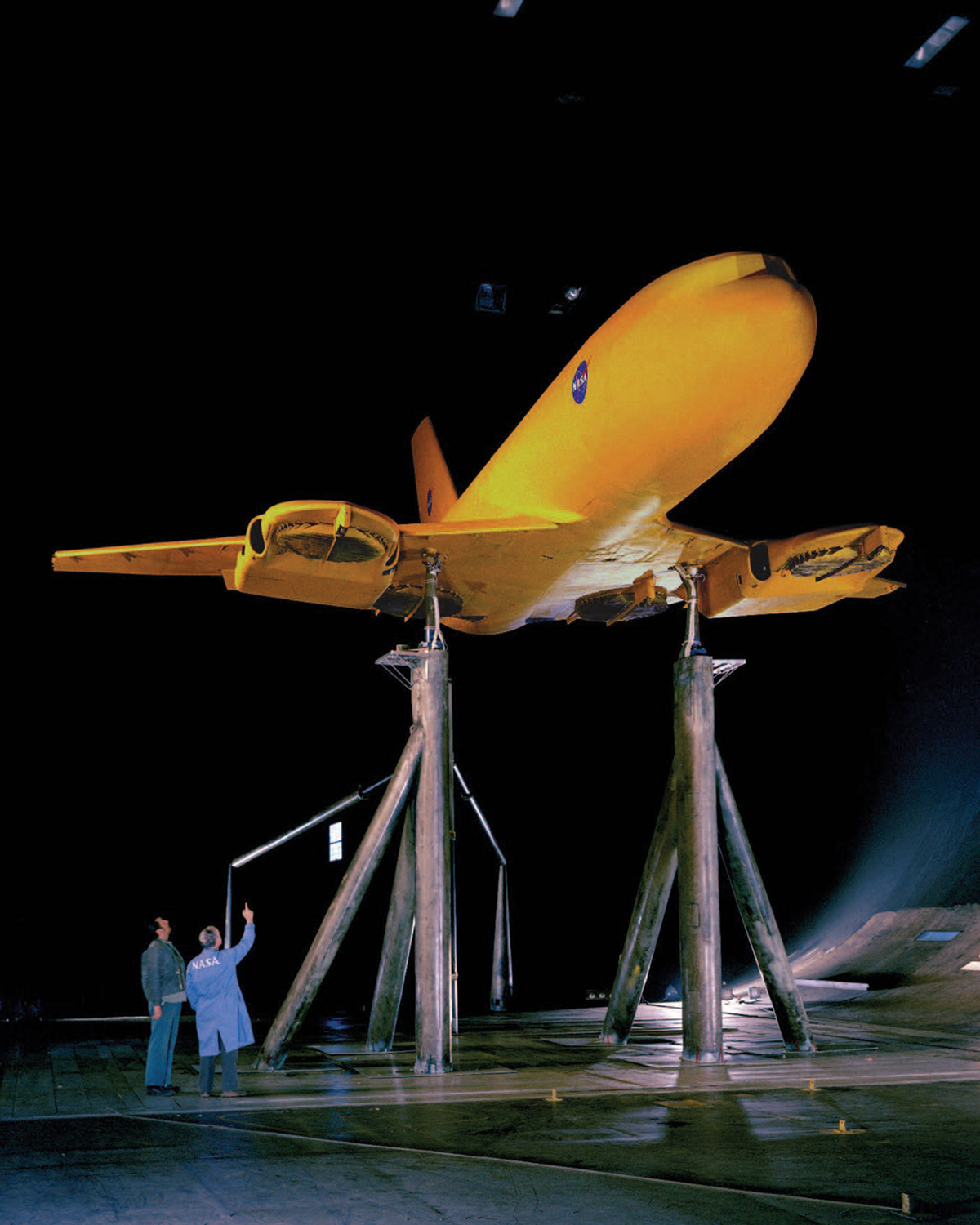 Two men look up at a yellow aircraft with a thin fan element attached to each wing, which is raised above the floor on tall columns inside a wind tunnel.