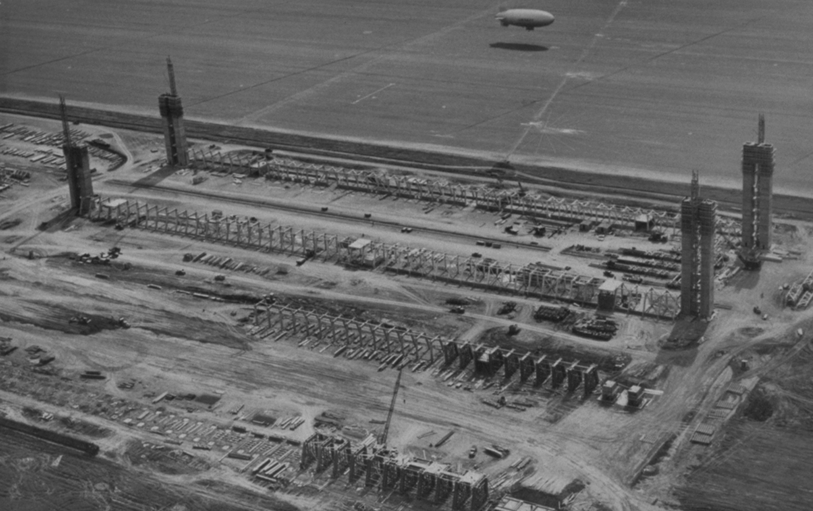 A birds-eye view of the early phase of construction for 2 blimp hangars located side-by-side. 4 concrete door towers rise from the Hangar 2 site.