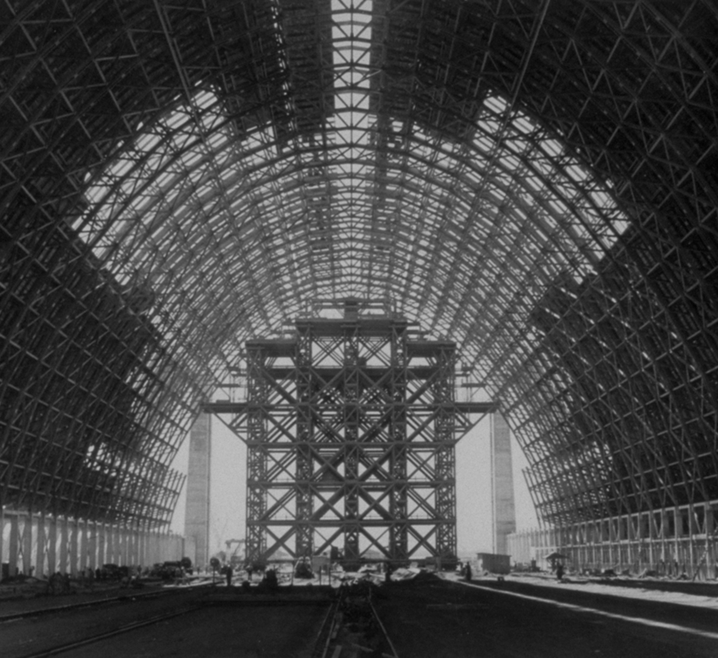 An interior view of the partially covered wood trusses of Hangar 2. A large scaffolding fills the space below the arches at the end of the building.