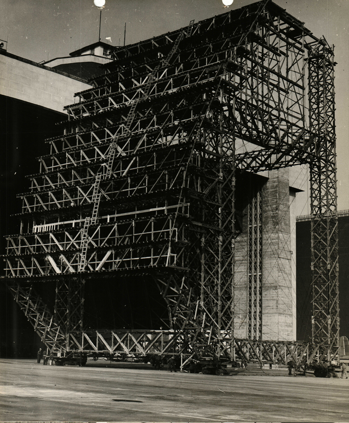 A tall, wide scaffolding tower on wheels waiting to be rolled into Hangar 3. It has a curved side that matches the parabolic arch trusses of the hangar.