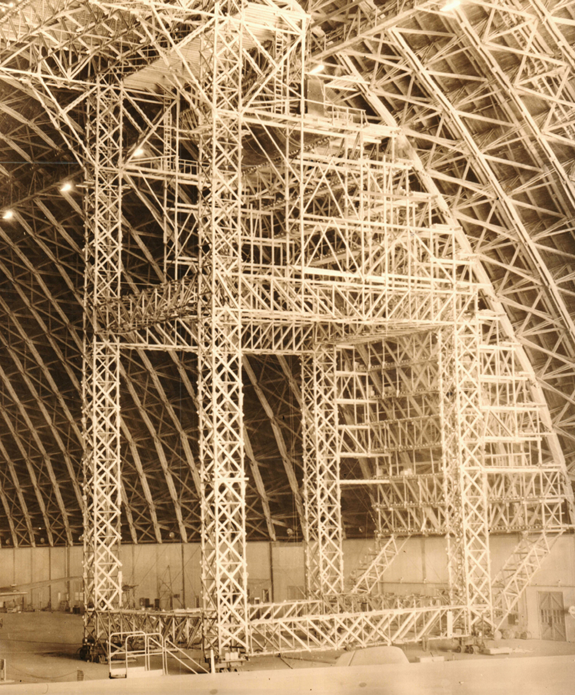 A tall, wide scaffolding tower inside the hangar. Its curved side mirrors the parabolic arch trusses of the hangar to efficiently fix the structure.