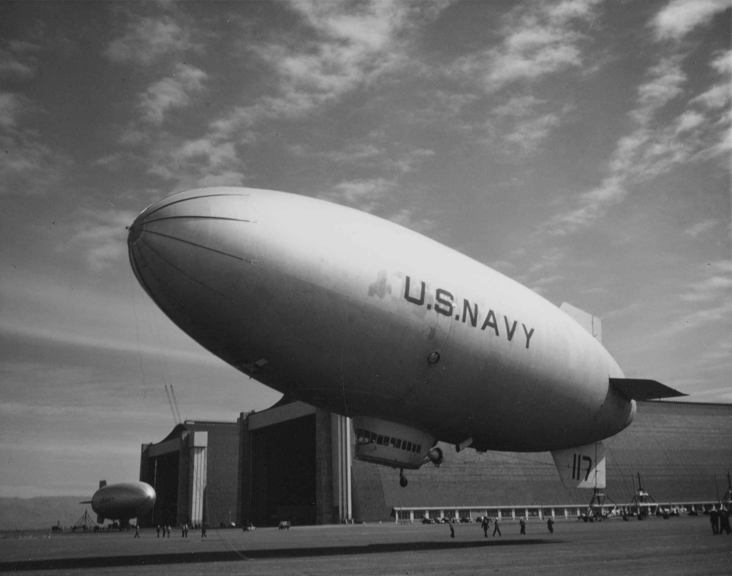 A blimp, with U.S. NAVY written on its side, lifts off near the open doors of Hangar 2. Ground crews prepare a blimp outside Hangar 3 for its next flight.