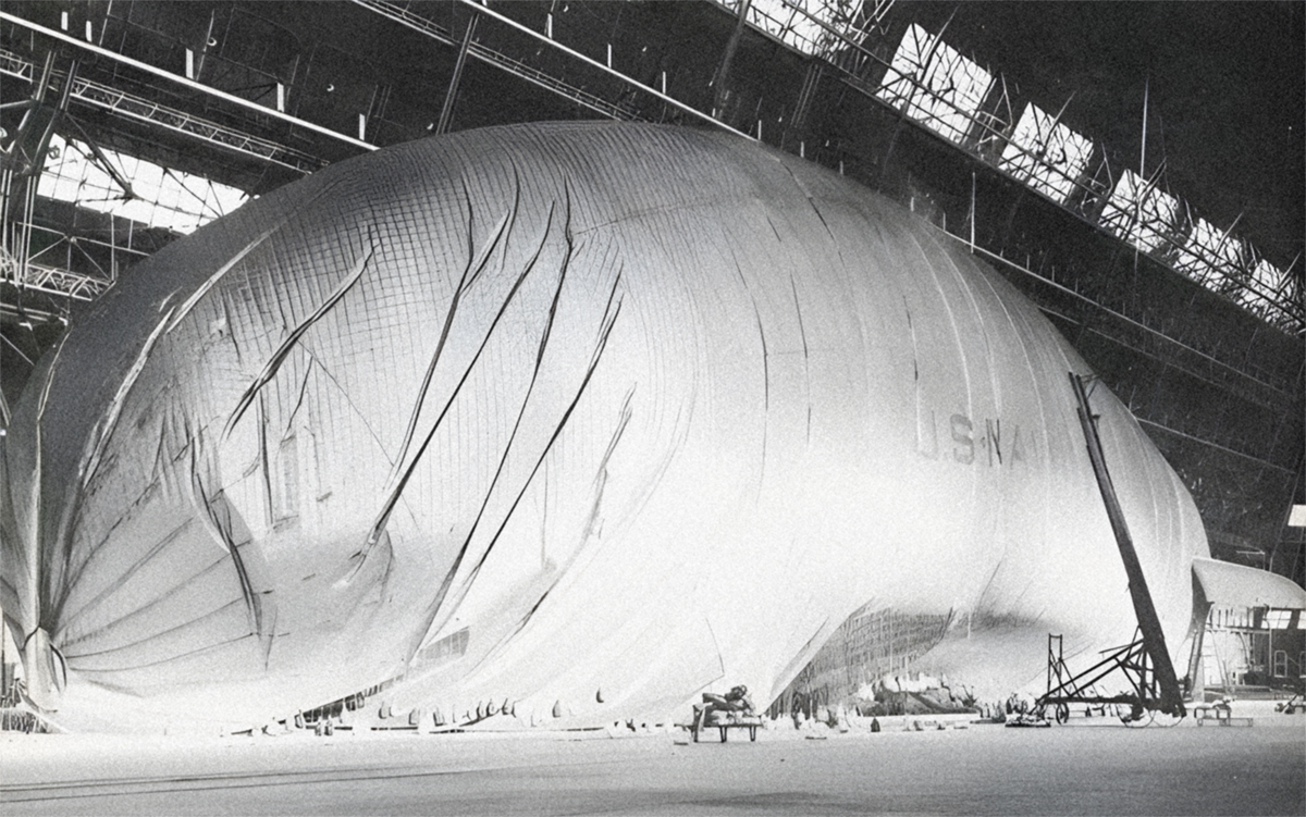 A partially deflated blimp, with faint U.S.NAVY lettering, begins to sag and crumple to the floor of Hangar 1. A crane is positioned beside the blimp.