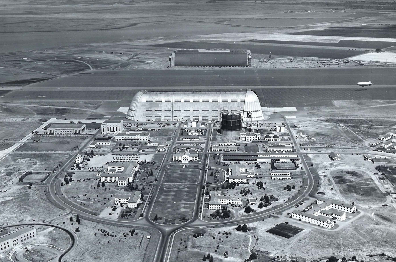 A birds-eye view of Hangar 1 and other buildings organized around a central lawn. A blimp takes off from the runway between Hangar 1 and Hangars 2 and 3.