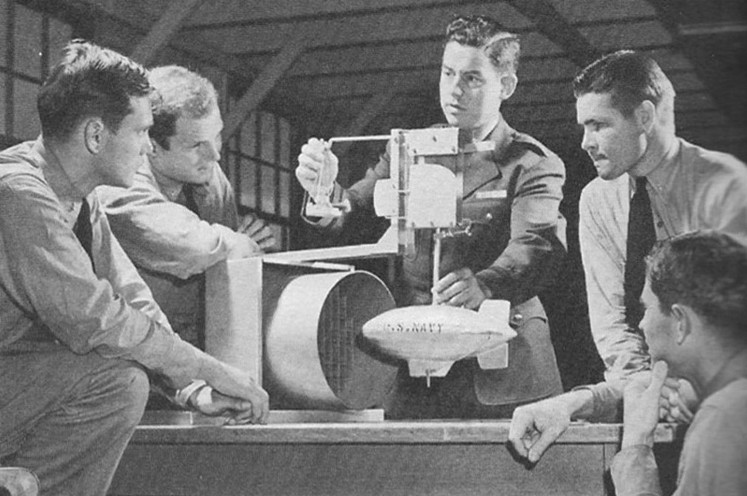 A Navy instructor holds two small posts attached to a tabletop model of a blimp near a fan. 4 cadets, on either side of their teacher, look at the model.