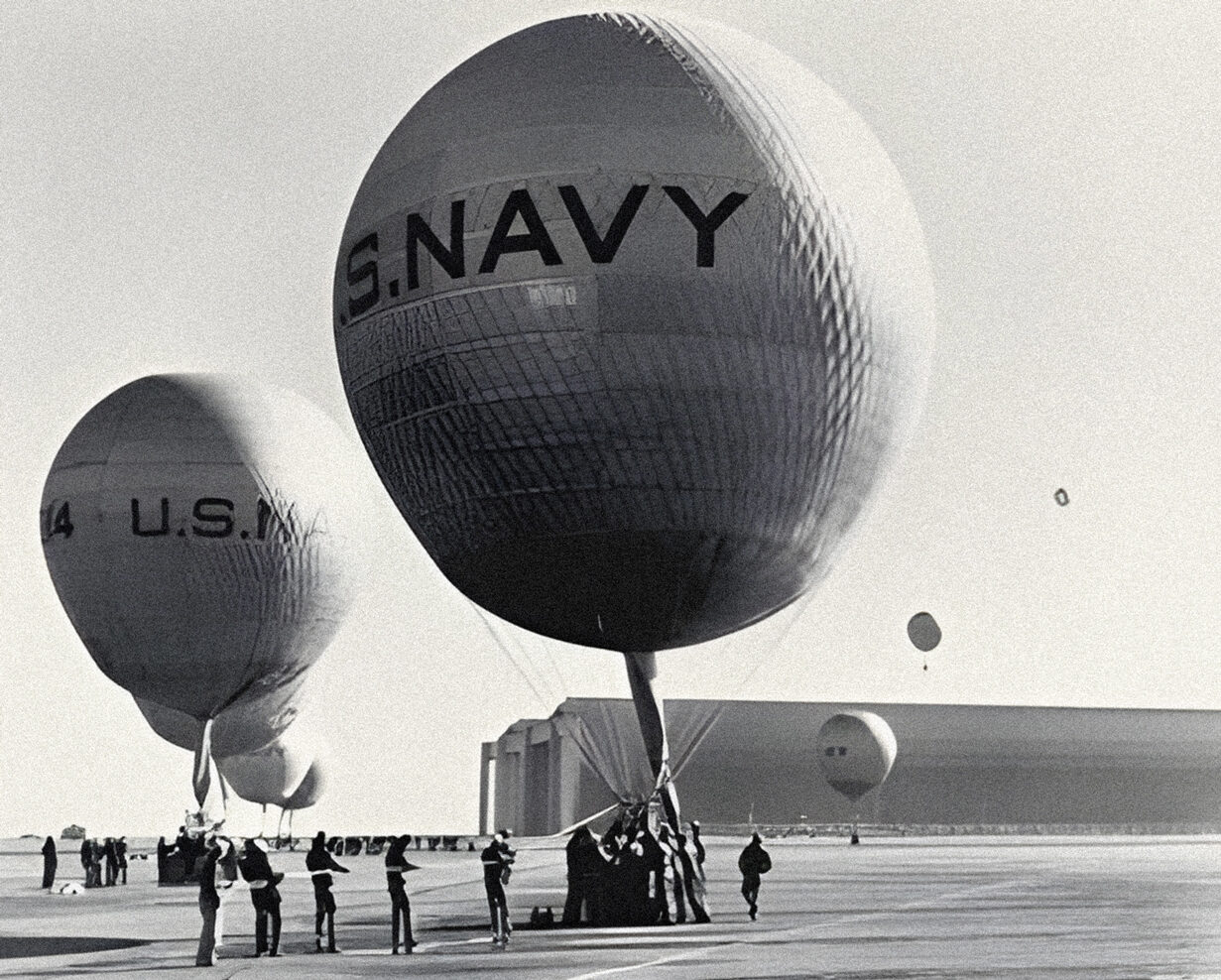 Ground crews prepare 6 large balloons, with U.S.NAVY written on their surfaces, for flight. In the distance, 2 balloons soar above Hangars 2 and 3.
