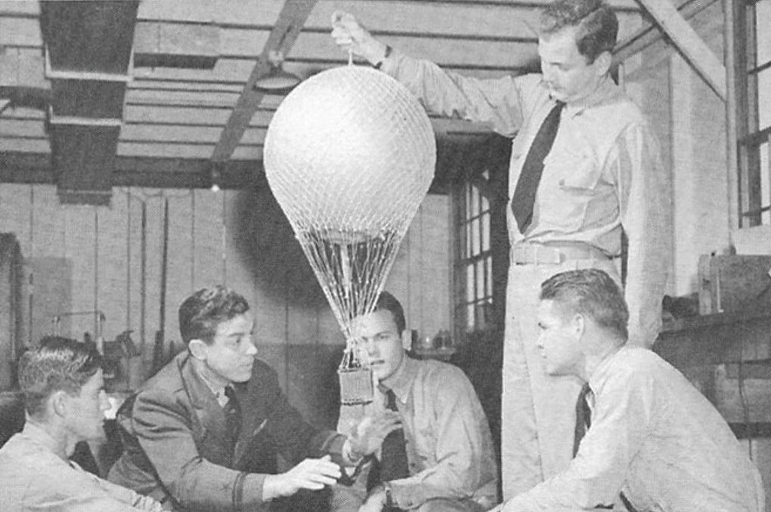 A Navy cadet holds the top of a training balloon model with his outstretched arm. The instructor, kneeling beside the model, teaches his 4 students