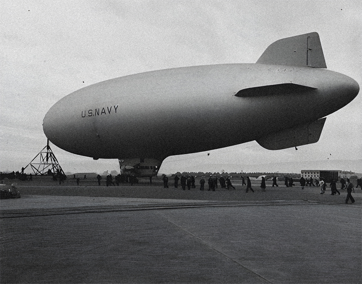 A blimp, with U.S.NAVY written on its side, is prepared for flight by a large ground crew walking under the blimp and near its triangular mooring mast.