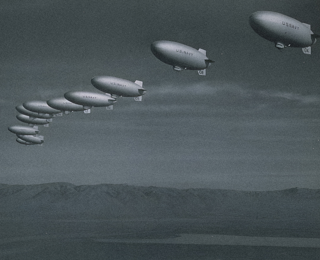 A curving row of 11 closely spaced blimps, with U.S.NAVY written on their sides, soar above a mountain ridgeline and the San Francisco Bay.