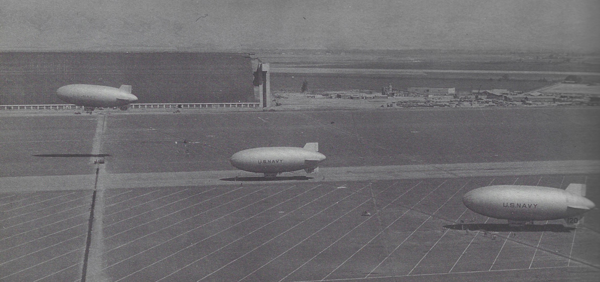 A diagonal row of 3 blimps, with U.S.NAVY written on their sides, on the runway beside Hangar 2. Two blimps wait to fly as the lead blimp takes off.