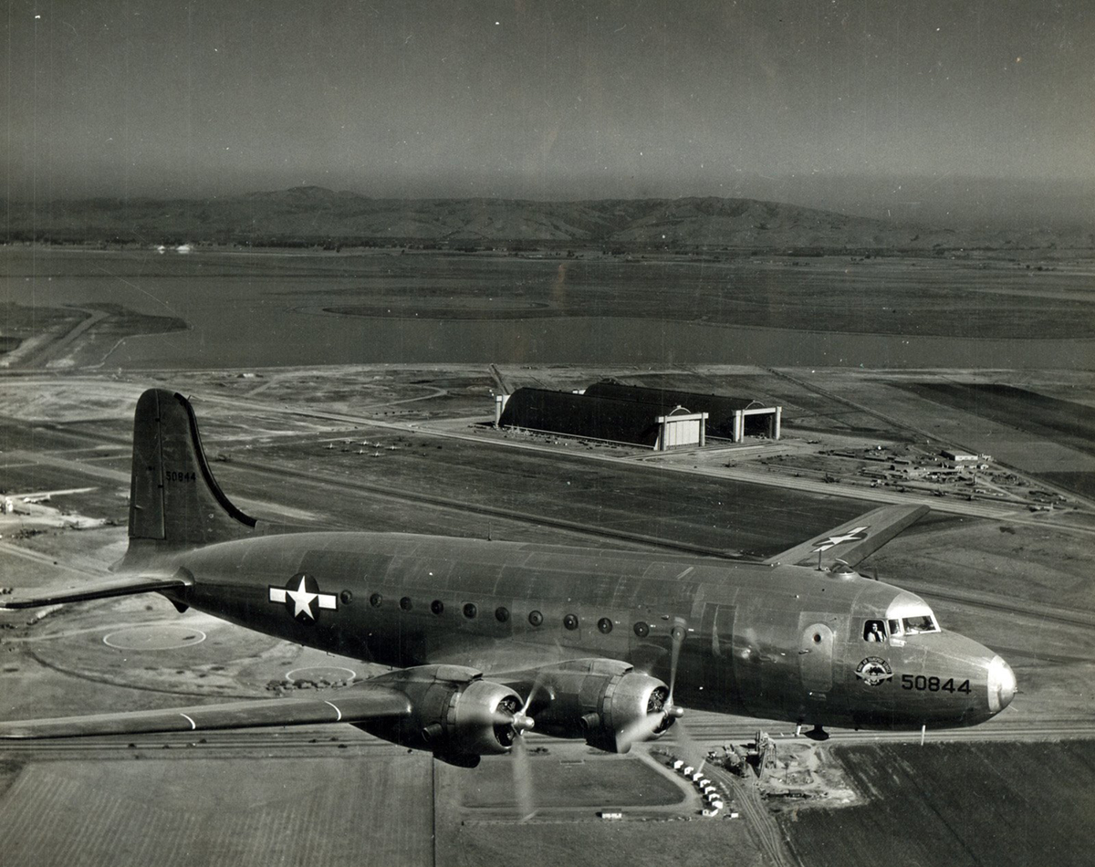 A side view of a 4-propeller aircraft, with the Navy white star insignia on its side and wing, soaring above Hangars 2 and 3 at Moffett Field.