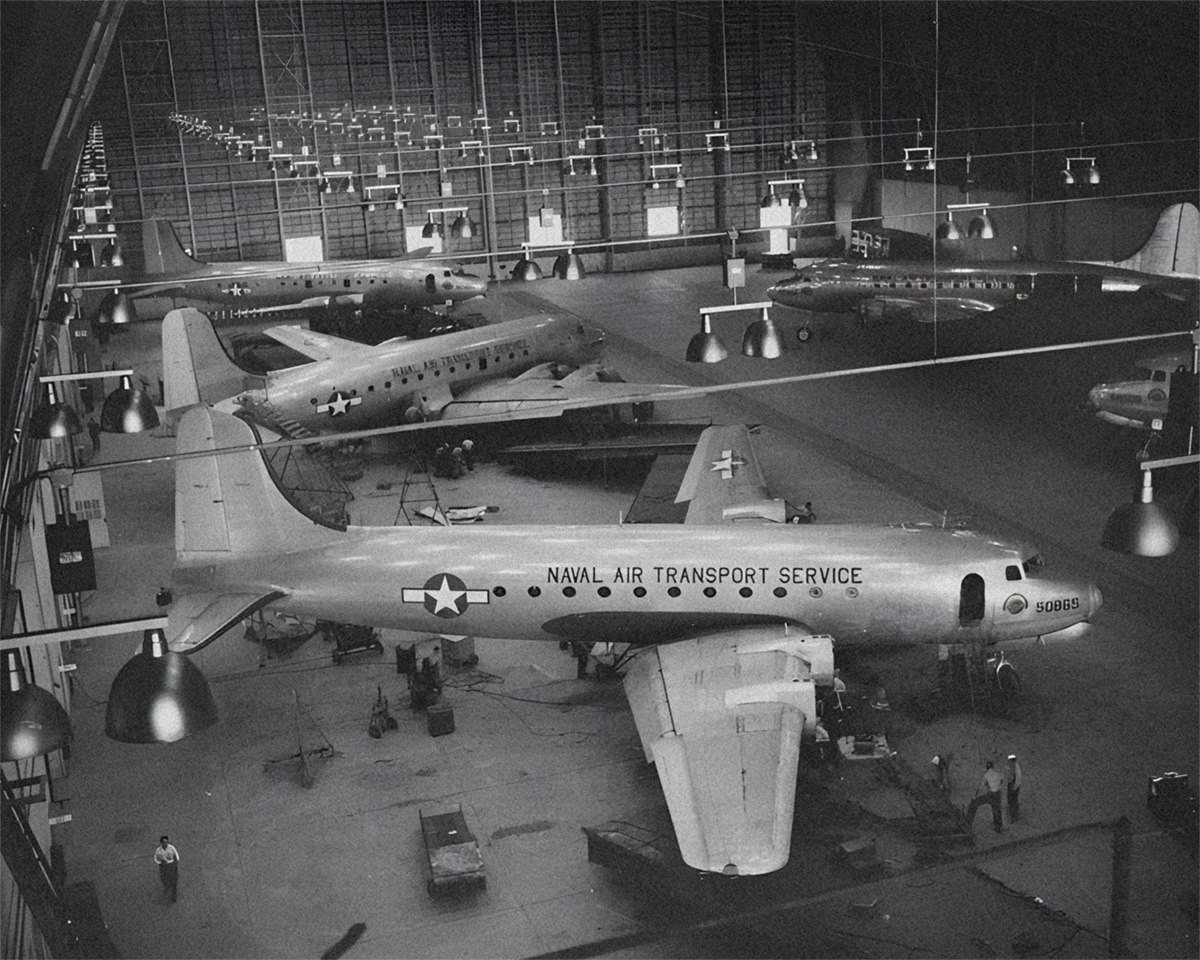5 aircraft inside a large maintenance hangar. Each plane has 4 propellers, a white star insignia and Naval Air Transport Service written on its side.