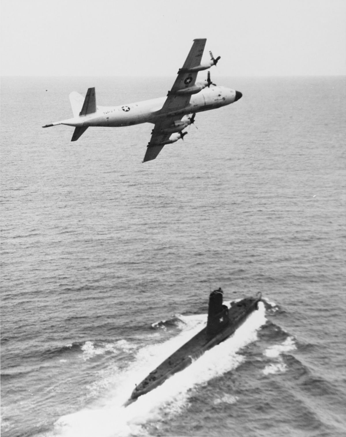A low-flying aircraft, with 4 propellers and the Navy white star insignia on its side and wing, tracks a dark submarine emerging from the ocean waves.