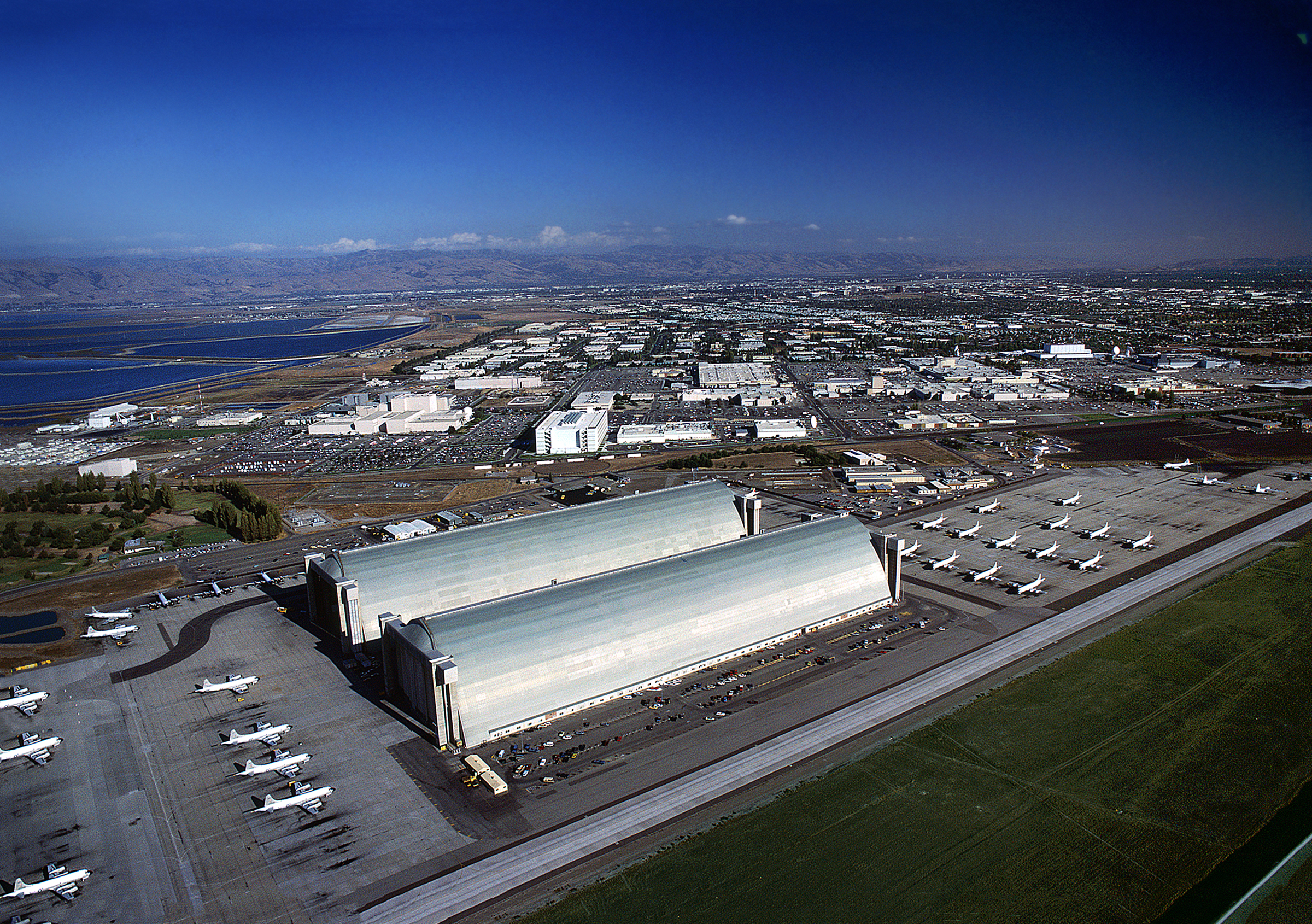 A birds-eye view of Hangars 2 and 3 located side-by-side at Moffett Field. Rows of P-3 Orion patrol planes are parked outside both ends of the hangars.